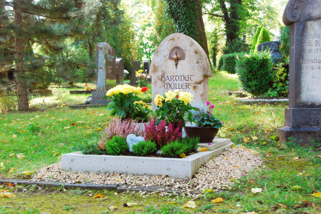 Aufwendige Grabgestaltung für den Herbst mit kleinen Büschen und Ziergräsern, sowie gelben Rosen in Vasen.