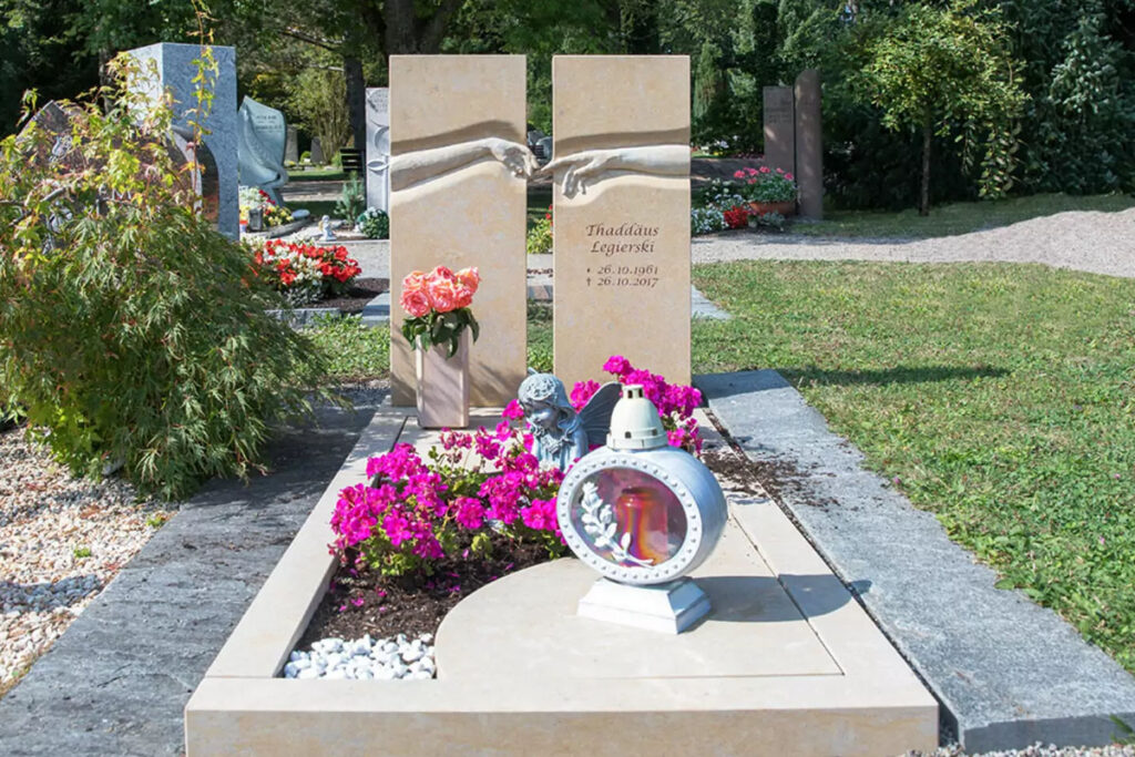  Ein geteiltes Grabmal mit berührenden Händen aus beigem Marmor, umgeben von einer Grabeinfassung und einem Bepflanzungsstreifen als Beispiel für eine Sommergrabgestaltung.