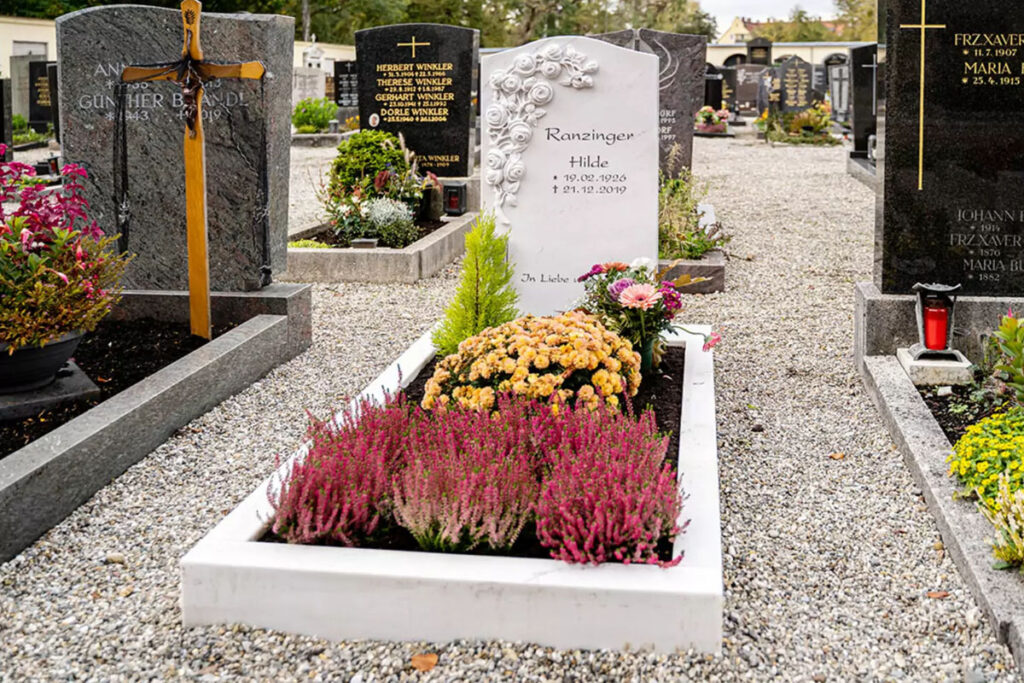 Ein klassischer Grabstein mit einem Rosenornament, umgeben von einer Grabeinfassung aus weißem Marmor sowie Grabgestaltung mit verschiedenen blühenden Pflanzen.