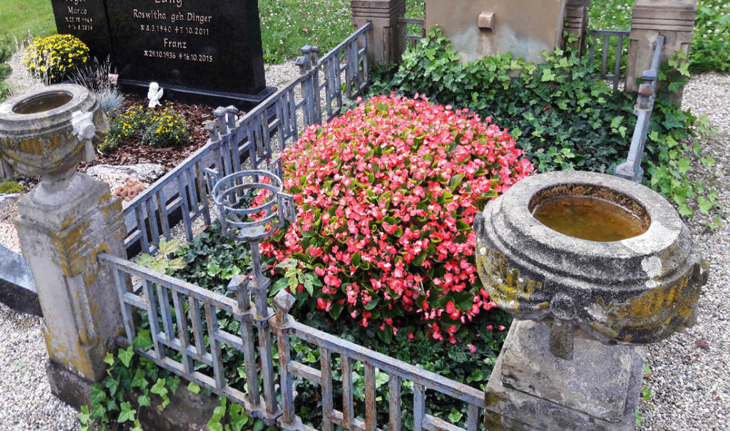 Kleines bepflanztes Familiengrab mit einer massiven Grabumrandung aus Stein und Zierzaun. Vorn am Zaun ist sogar eine Halterung für eine Grablampe.