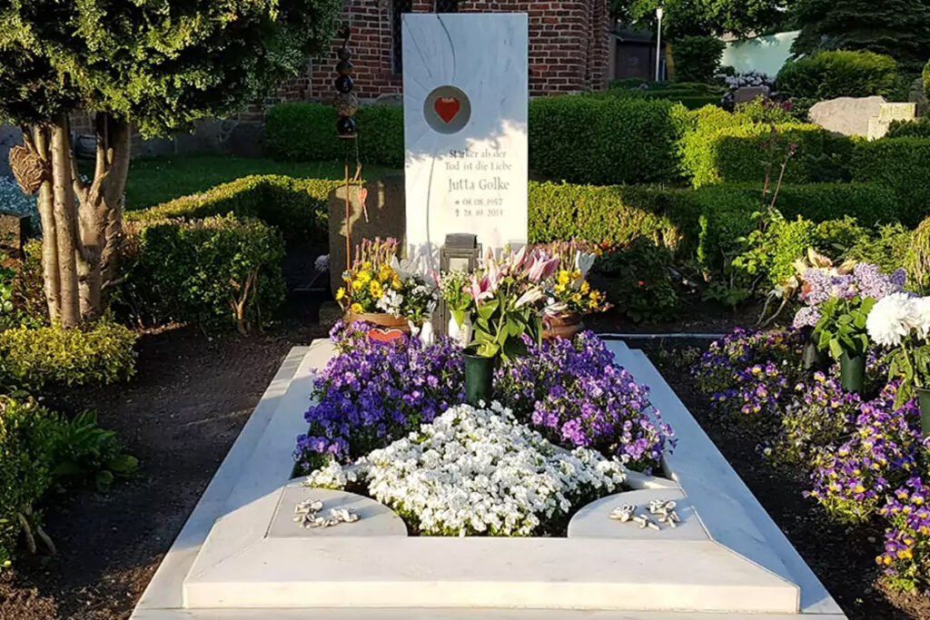 Ein Grabstein mit einem Glaselement in Herzform für ein Einzelgrab aus Marmor mit Einfassung und Grabplatten, umgeben von einer modernen Grabbepflanzung im Sommer mit vielen verschiedenen Blumenarten.
