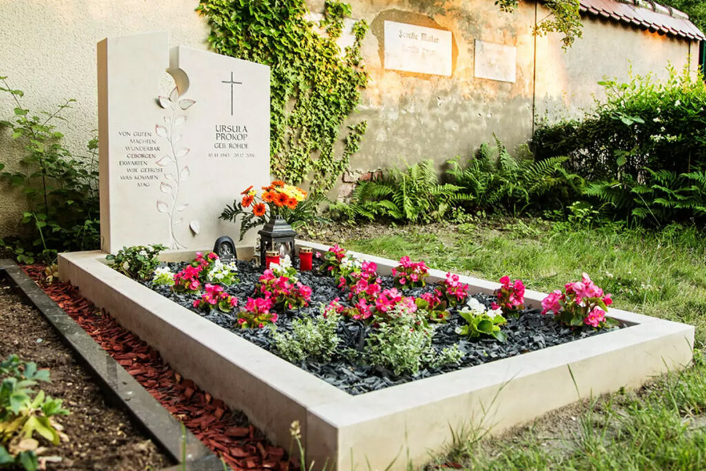 Ein moderner Einzelgrabstein aus Kalkstein, umgeben von einer passenden Einfassung und pflegeleichte Grabbepflanzung mit bunten Blumen und Rindenmulch Untergrund.