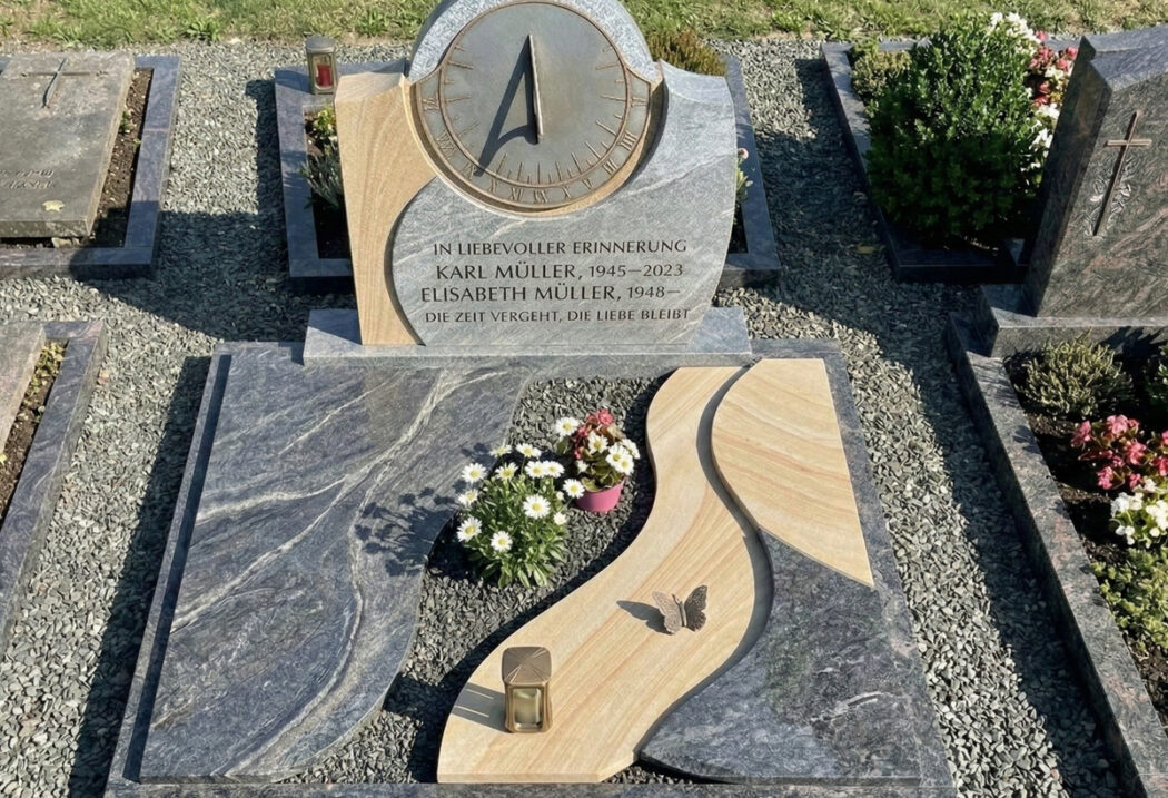 Diese moderne Grabanlage kombiniert kontrastierende Steine: dunklen  polierten Granit mit grauer Wellenmaserung und hellen Sandstein mit Schichtung. Hinzu kommen Bronzeelemente wie ein Schmetterling und eine Laterne sowie eine kreisrunde Metall-Sonnenuhr. Das Ensemble liegt auf feinem grauen Kies.