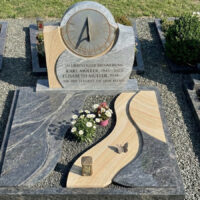 Diese moderne Grabanlage kombiniert kontrastierende Steine: dunklen  polierten Granit mit grauer Wellenmaserung und hellen Sandstein mit Schichtung. Hinzu kommen Bronzeelemente wie ein Schmetterling und eine Laterne sowie eine kreisrunde Metall-Sonnenuhr. Das Ensemble liegt auf feinem grauen Kies.