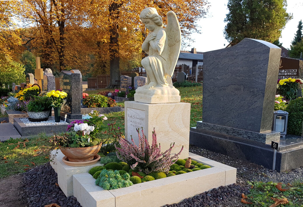 Dieses stimmungsvolle Bild zeigt eine liebevoll gestaltete Grabstätte im warmen Licht der Herbstsonne. Im Zentrum steht eine knieende Engelsfigur aus hellem Sandstein  die auf einem quadratischen Sockel thront. Der Sockel trägt die Inschrift für Ruth Schmiedel.