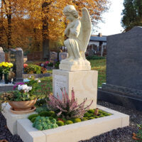 Dieses stimmungsvolle Bild zeigt eine liebevoll gestaltete Grabstätte im warmen Licht der Herbstsonne. Im Zentrum steht eine knieende Engelsfigur aus hellem Sandstein  die auf einem quadratischen Sockel thront. Der Sockel trägt die Inschrift für Ruth Schmiedel.