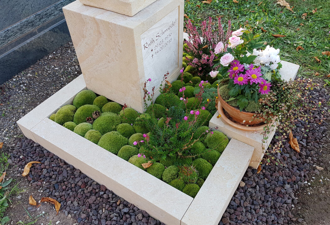 Das Grab wird durch eine massive  helle Steineinfassung in L-Form begrenzt  die sich deutlich von dem dunklen Lavakies der unmittelbaren Umgebung abhebt. Diese klare Abgrenzung verleiht der Ruhestätte eine geordnete Struktur und lässt die Pflanzen sowie den hellen Stein leuchten.