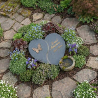 Im Zentrum ruht ein zweigeteilter  anthrazitfarbener Grabstein in Herzform für Ernst Sempera  verziert mit einem filigranen Bronze-Schmetterling.