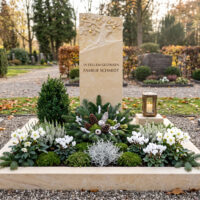 Grabgestaltung zu Allerheiligen mit schöner Deko schlichter Einzelgrabstein aus Sandstein mit einem gravierten Lebensbaum und Vergoldungen einschließlich Umrandung und moderner Grablampe aus Bronze mit einer Bepflanzung zu Allerheiligen.